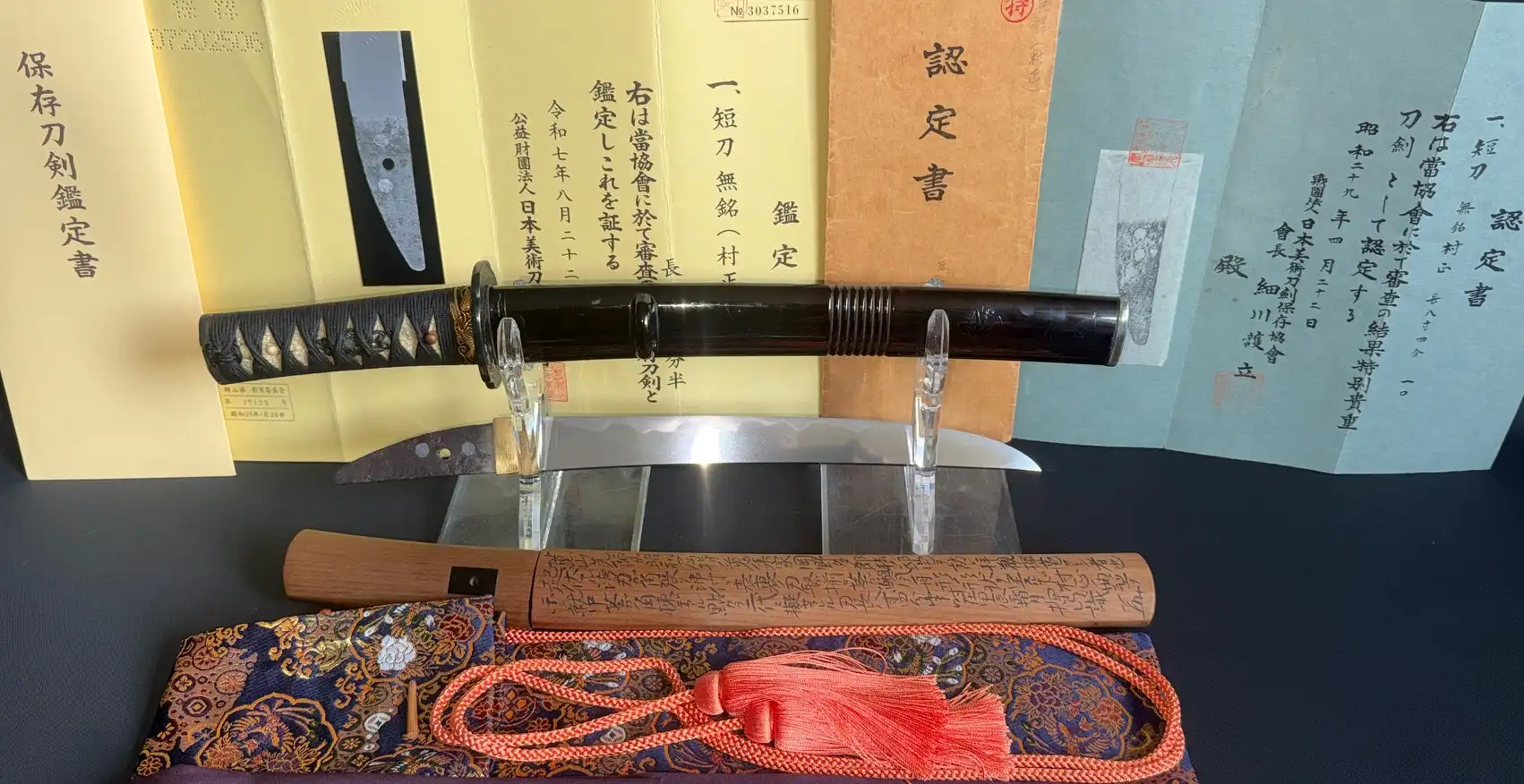 Nidai Muramasa Sayagaki katana with dark scabbard resting on a stand, paper certificates behind it