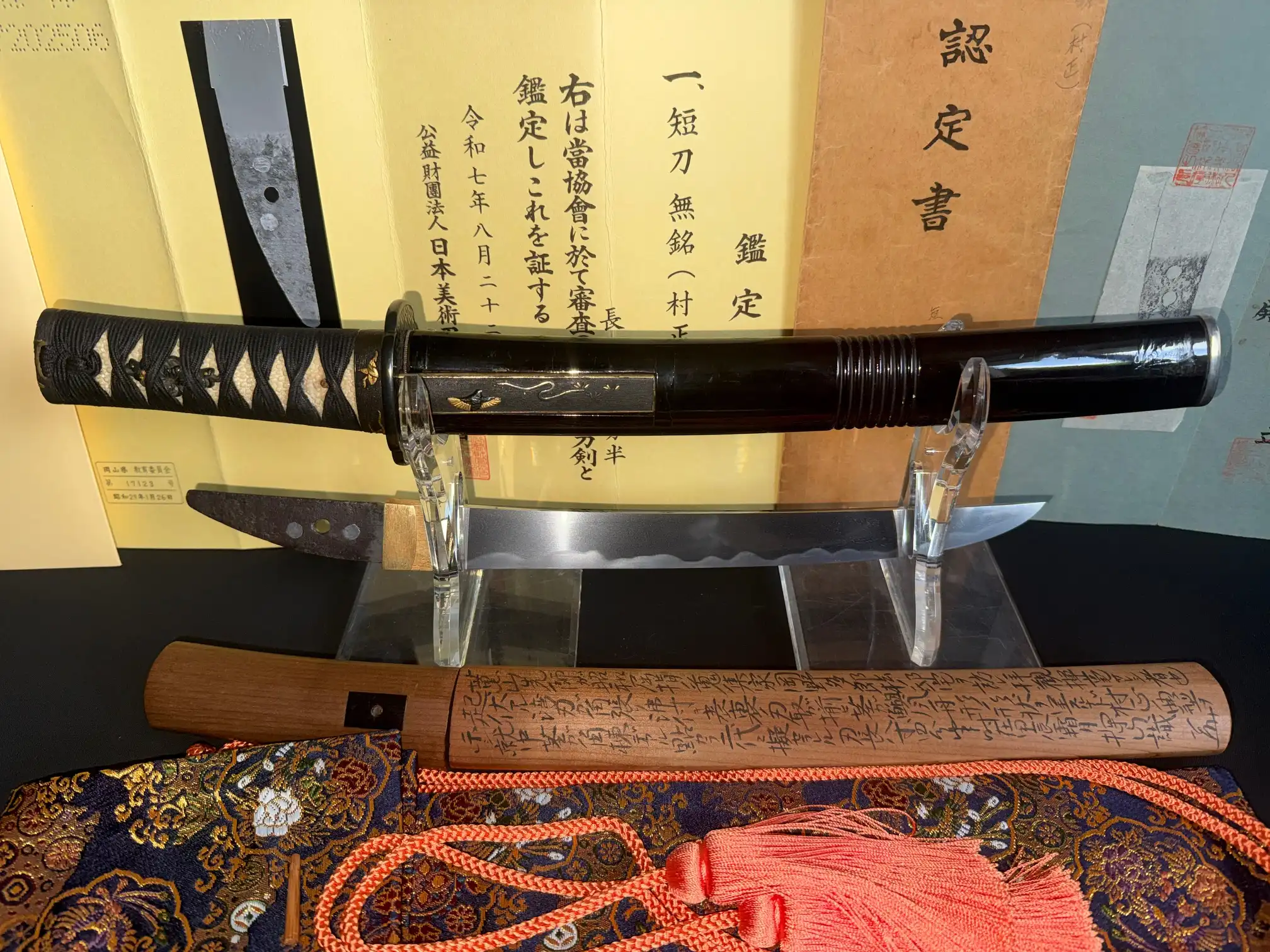 Nidai Muramasa Sayagaki katana, blade visible on a stand with paperwork, pommel and wooden scabbard below