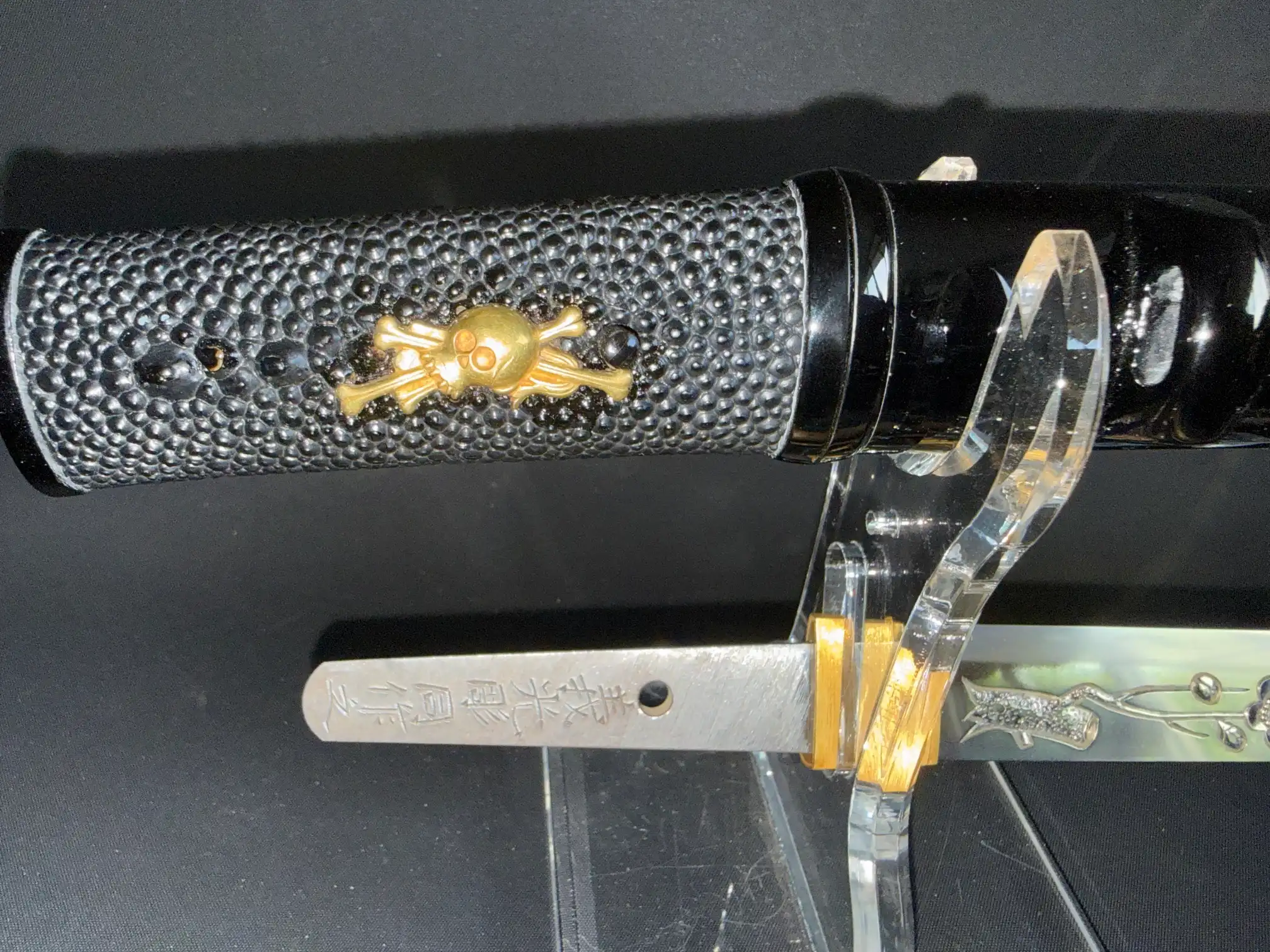 Ōno Yoshimitsu Mukansa Tantō with black ray skin handle and gold skull insignia, displayed on a stand.