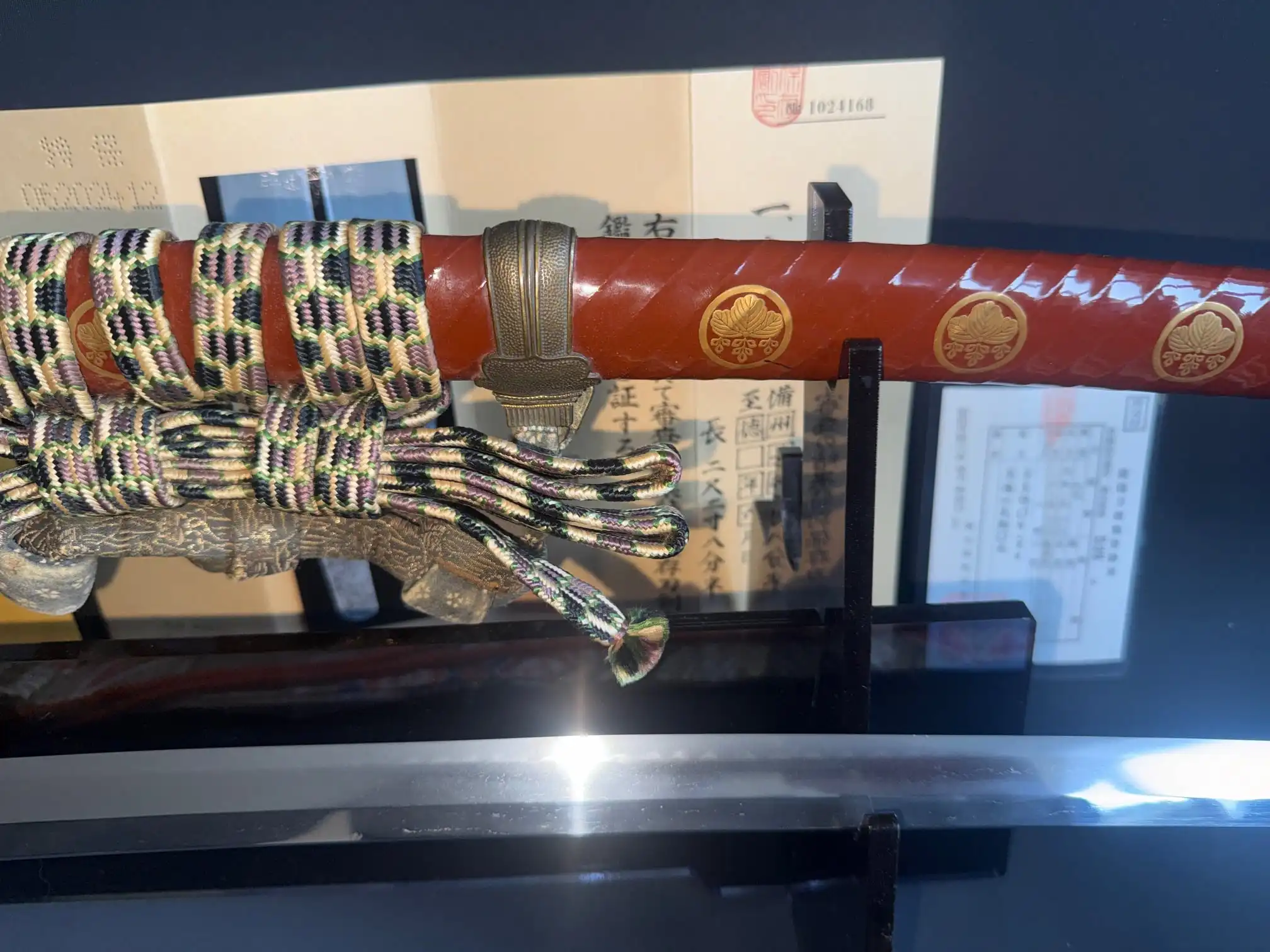 Close-up of a Nanbokucho Tachi Hidemitsu katana showing the red saya, gold mon, and intricate tsuka-ito
