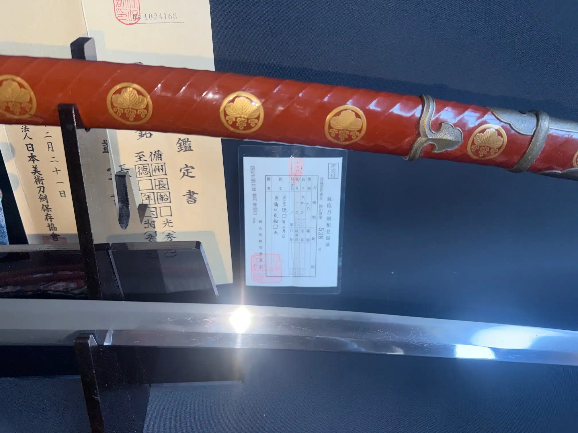 Close-up of a Nanbokuchō Tachi Hidemitsu katana with a red saya ornate with golden emblems, and its blade