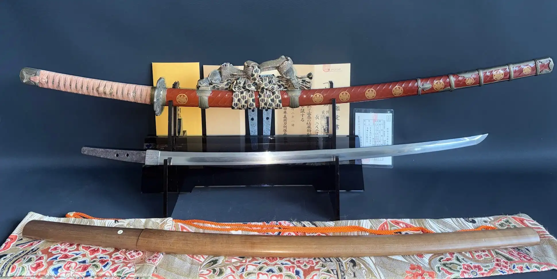 Full view of a red and gold Nanbokuchō Tachi Hidemitsu katana with white tsuka, paperwork, and white scabbard.