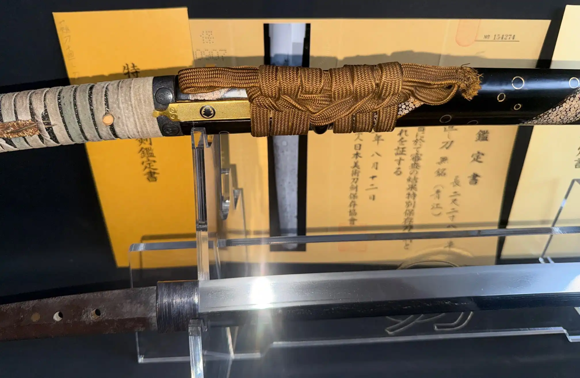 Close-up of an Aoe Nagamaki-Naoshi Katana blade and handle with golden Ito wrap, certificates in background.