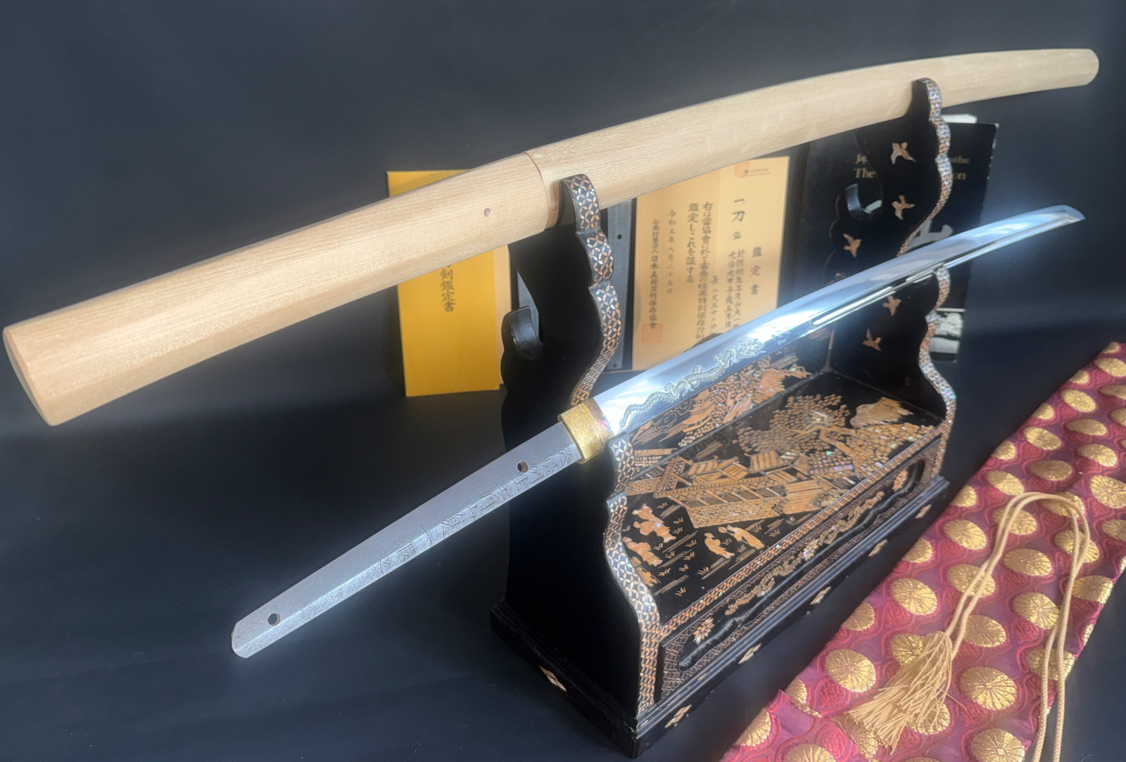 Japanese Masterpiece Katana by Gassan Sadakazu, blade displayed on stand with shirasaya and documentation in background.