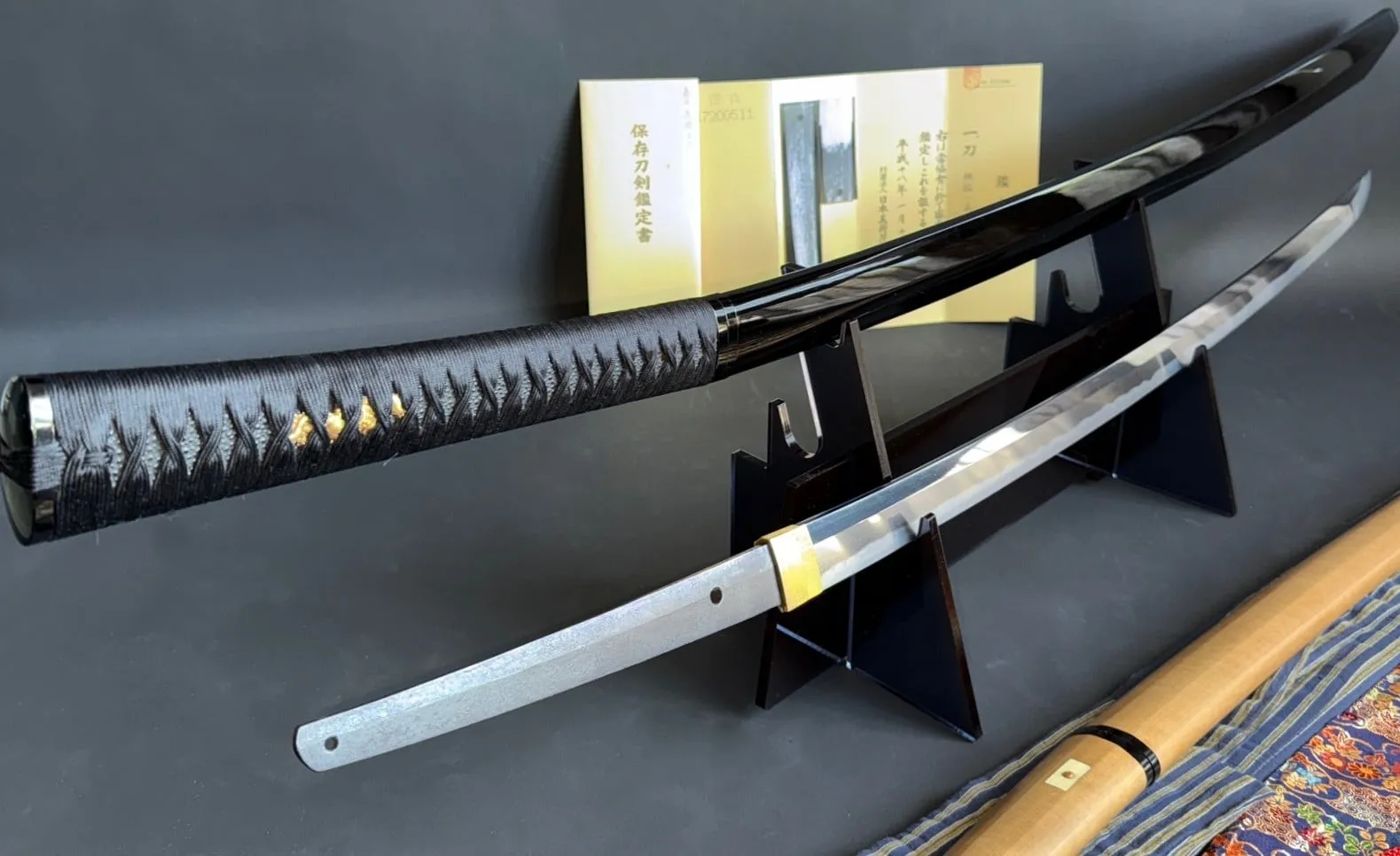 Jinja Hōnō Tachi Naganobu katana with black tsuka, visible gold under wrap, and polished blade on stand