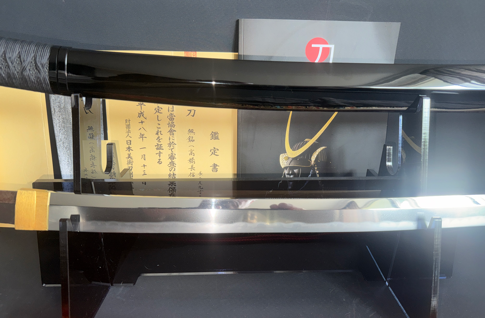 Jinja Hōnō Tachi Naganobu Katana display with black lacquered saya and polished blade, Japanese text behind