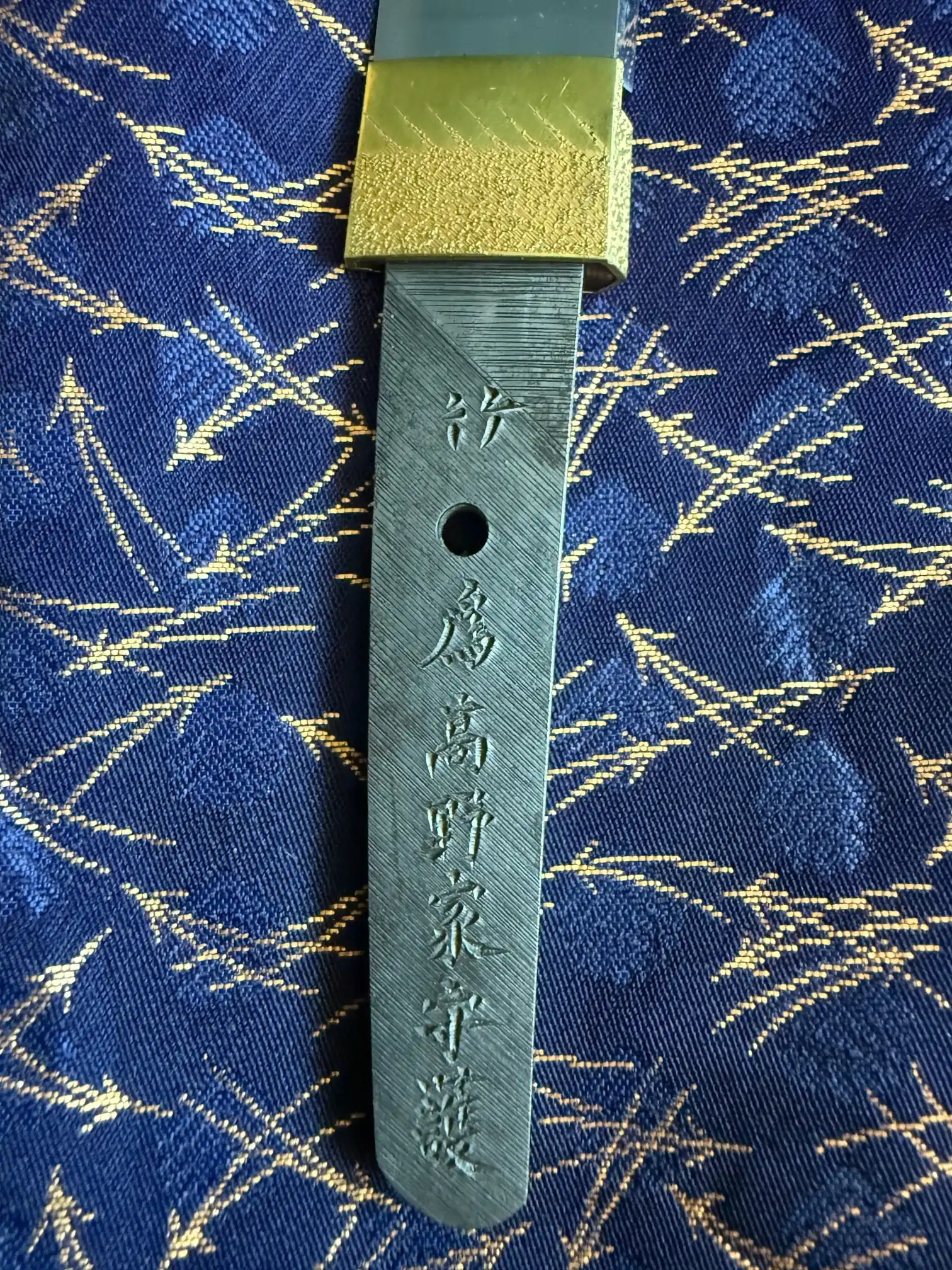 Close-up of the engraved tang of the Tanto Gassan Sadakatsu, with a golden habaki and blue fabric in the background
